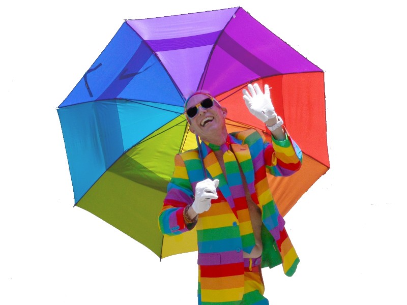 Laughing person with their head tossed back. They are wearing sunglasses and clothing with wide rainbow-coloured stripes. Their right hand is holding a large rainbow-coloured umbrella that is balanced on their right holder.