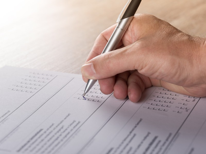 Close-up photo of a right hand holding a pen and filling out a paper form with tick boxes.