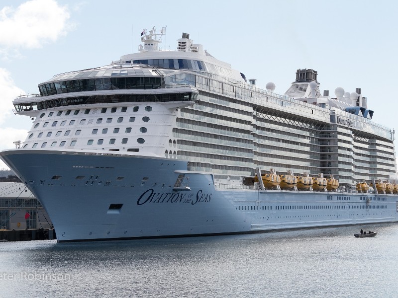 A massive white cruise ship anchored at a dock with its stern towards the camera. The words ‘Ovation of the Seas’ are faintly visible on the ship, which dwarfs a boat on the right.