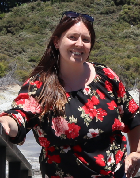 Katie Haggath standing outside in the sun with trees in the background and her hand on a railing. She's wearing a black and red floral top and is smiling at the camera