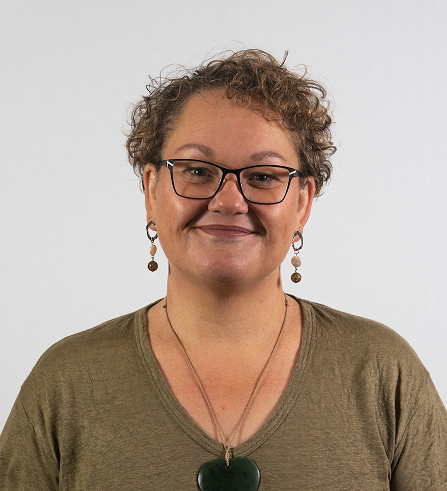 A headshot of Kylee smiling - she has short, curly brown hair and is wearing glasses, earrings, and a pounamu carving necklace