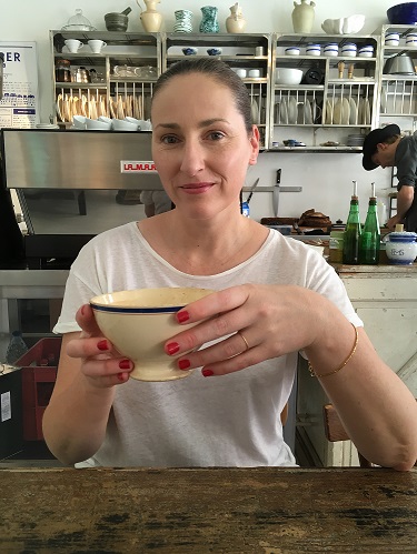 A photo of Jody smiling - she has hair hair pulled back, is sitting at a table holding a white mug of coffee and is wearing bright red nail polish.